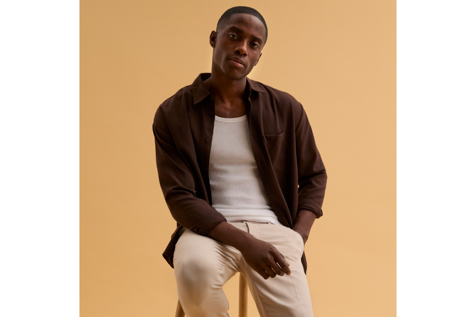 A man wearing an unbuttoned brown shirt over a cream top and trousers from New Look
