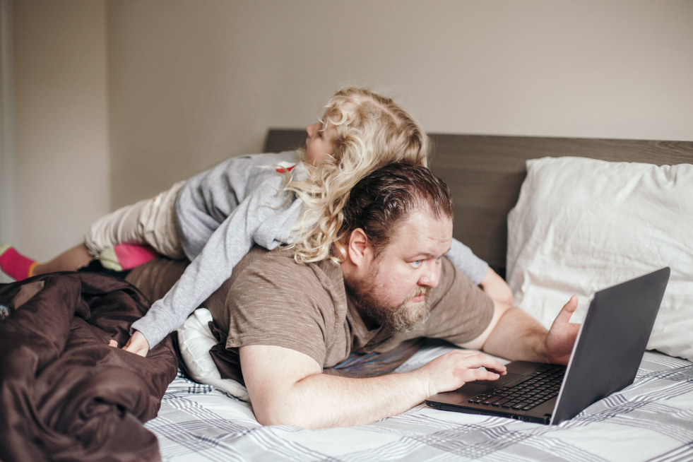 A dad trying to work on a laptop while his daughter lies on his back
