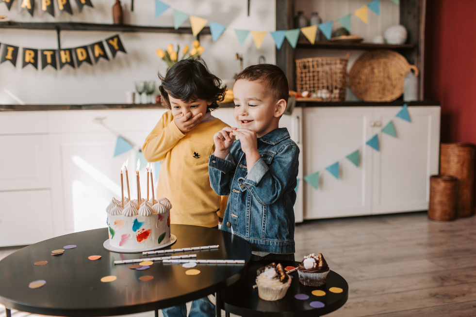Two young boys stood next to a birthday cake in a kitchen 