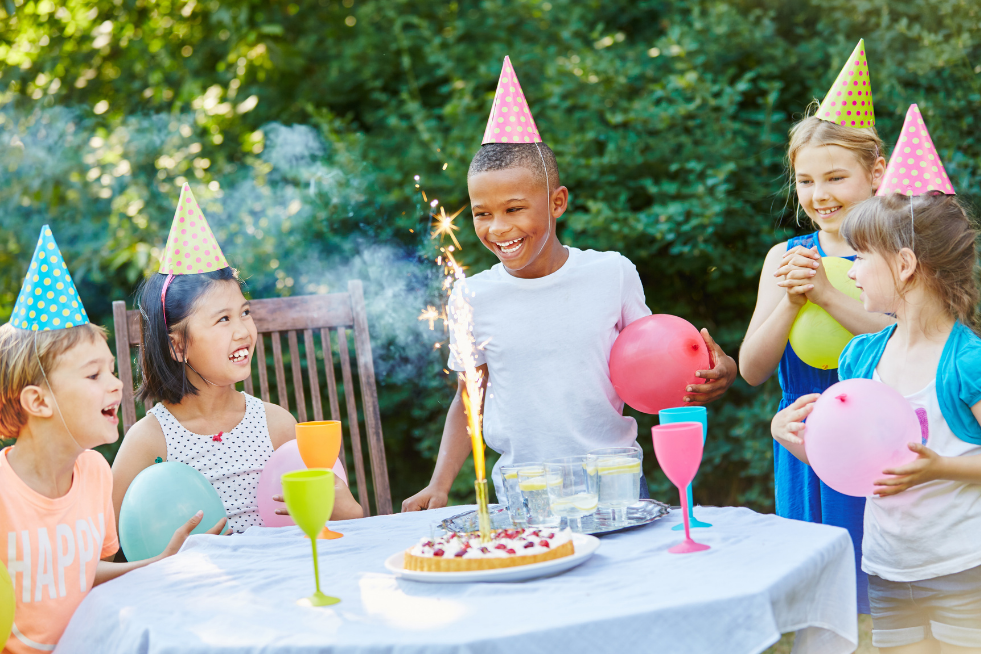 A group of children with party hats on and a cake with a sparkler 
