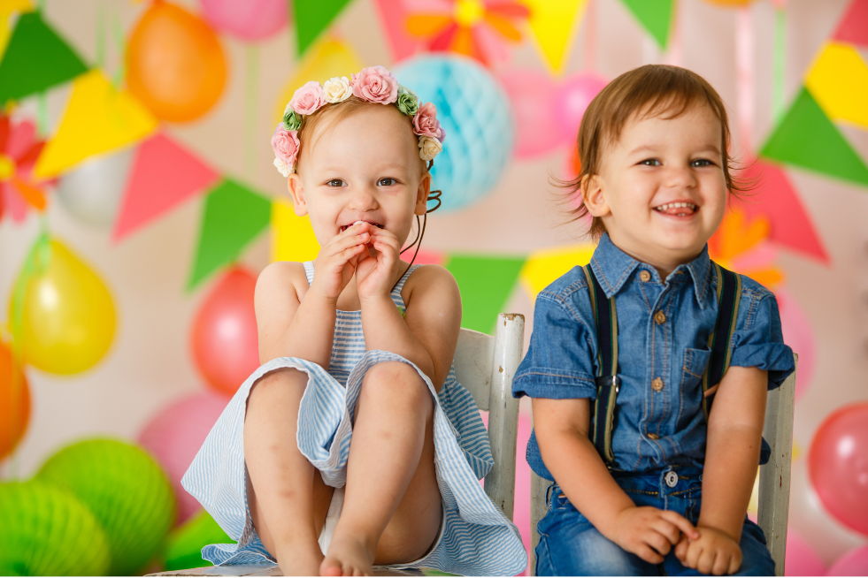 Two small children smiling at the camera with bunting and balloons in the background